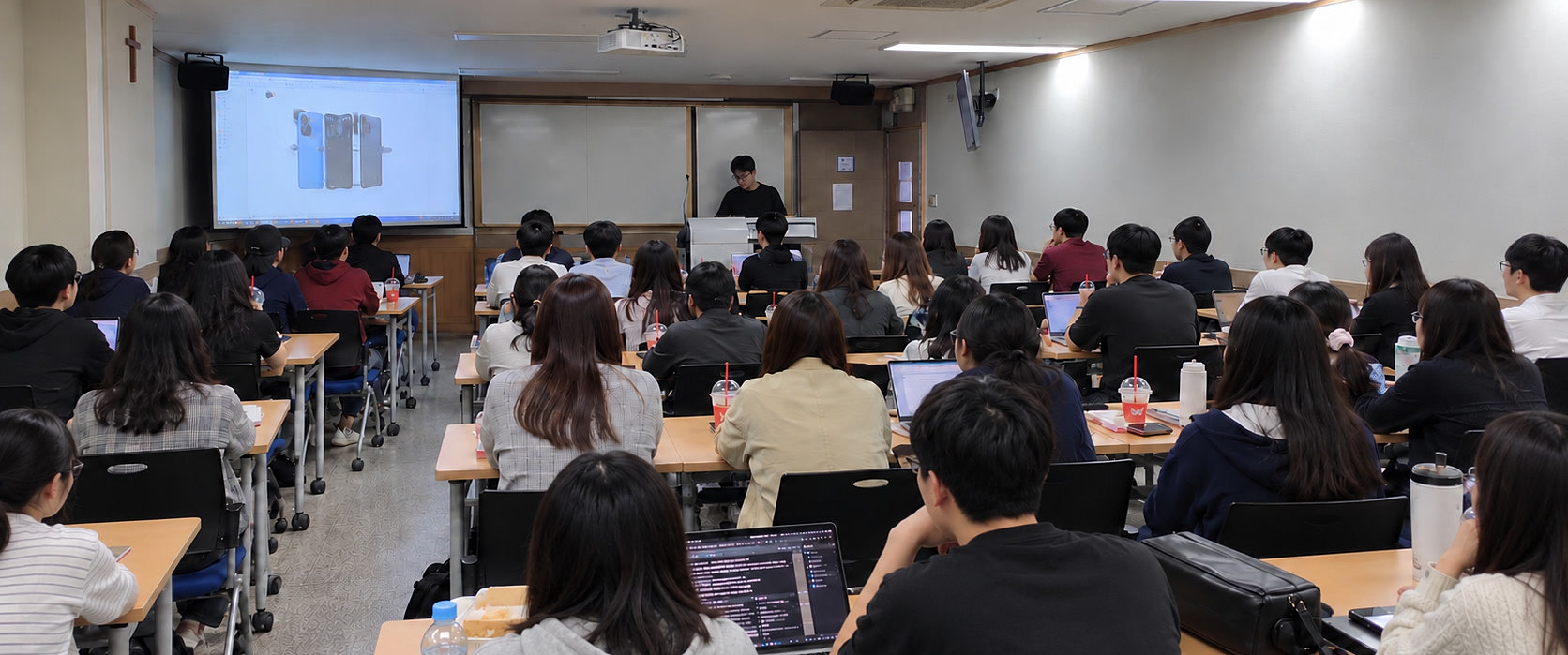 대학 실습 교육 현장 — 수강생들이 노트북으로 실습하고 강사가 발표 화면으로 설명하고 있다